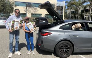 Two people displaying cat and dog food donations outside a car with the a white dog leaning out a window that is rolled down
