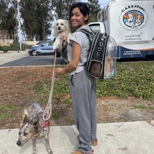 Girl at C.A.R.E.4Paws Mobile Pet Wellness Clinic with two dogs, one on lead and one in her arms. She is also wearing a cat backpack with a cat inside.